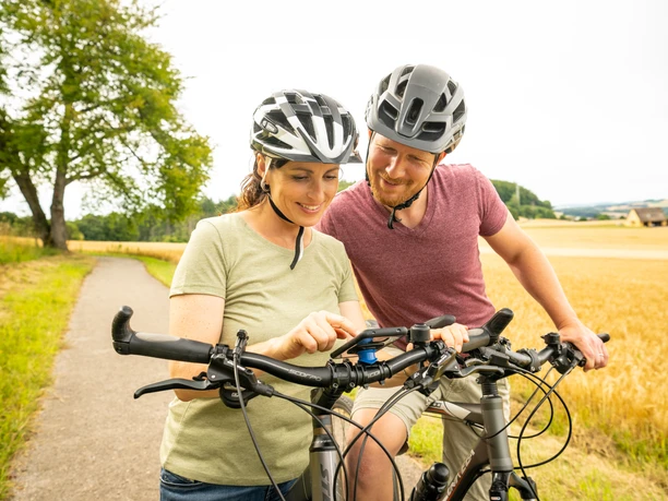 Eine lächelnde Frau und ein Mann in Fahrradhelmen schauen auf ein GPS-Gerät befestigt an einem Fahrradlenker.