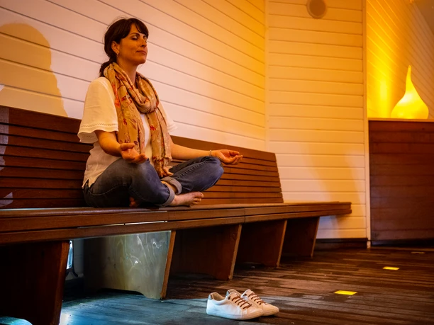 Bad Salzuflen-Gradierwerke Solekammer-Teutoburger-Wald-Tourismus-D-Ketz-056.jpg Eine Frau meditiert in einem ruhigen, warm beleuchteten Raum auf einer Holzbank, Schuhe neben ihr.