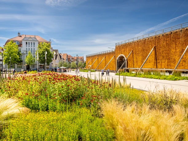 Gradierwerk mit Solewand in einer Parkanlage, umgeben von blühenden Pflanzen und einem klaren Himmel.