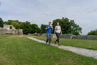 Bueren_BurgruineRingelstein_Teutoburger-Wald-Tourismus-Patrick-Gawandtka-096.jpg Ein Paar spaziert fröhlich auf einem Pfad durch eine grüne Wiese, mit Burgruine im Hintergrund.