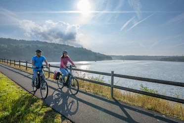 BadWuennenberg–Aabachtalsperre-Teutoburger-Wald-Tourismus-Patrick-Gawandtka-023.jpg Zwei Radfahrer fahren entspannt am Morgen entlang eines ruhigen Sees unter klarem, blauem Himmel.