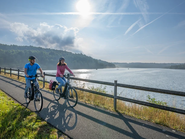 BadWuennenberg–Aabachtalsperre-Teutoburger-Wald-Tourismus-Patrick-Gawandtka-023.jpg Two cyclists ride relaxed in the morning along a calm lake under a clear blue sky.