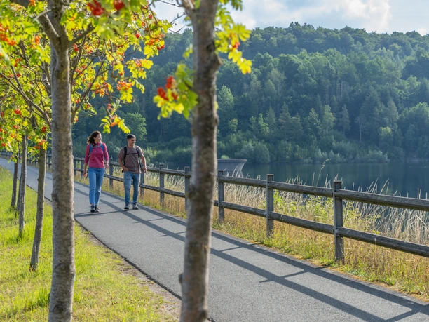 BadWuennenberg–Aabachtalsperre-Teutoburger-Wald-Tourismus-Patrick-Gawandtka-033.jpg Two people are walking along a lake, surrounded by green trees and a blue sky.
