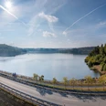 Stausee mit Radweg im Vordergrund, umgeben von dichtem, grünen Wald unter klarem blauen Himmel.Stuwmeer met fietspad op de voorgrond, omgeven door dicht, groen bos onder een strakblauwe hemel.Reservoir with cycle path in the foreground, surrounded by dense, green forest under a clear blue sky.