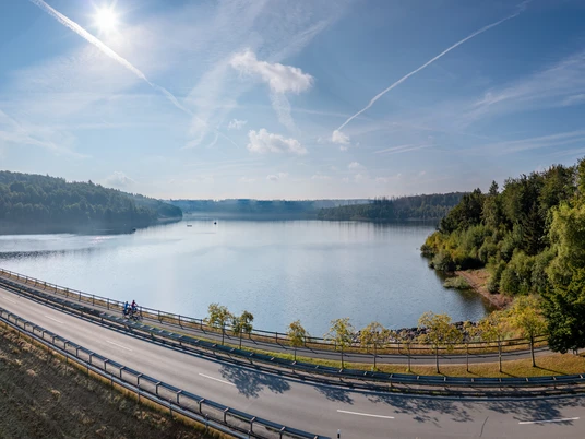 BadWuennenberg–Aabachtalsperre-Teutoburger-Wald-Tourismus-Patrick-Gawandtka-038-CC-BY-SA.jpg Stausee mit Radweg im Vordergrund, umgeben von dichtem, grünen Wald unter klarem blauen Himmel.