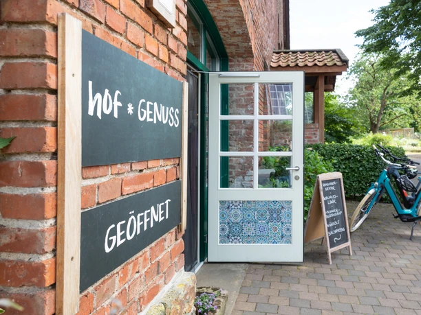 Steinheim-Hagedorn-Hofladen-Teutoburger-Wald-Tourismus-A-Röser-118.jpg Eingang eines Backsteingebäudes mit Schildern "hof * genuss" und "GEÖFFNET", daneben Fahrräder.