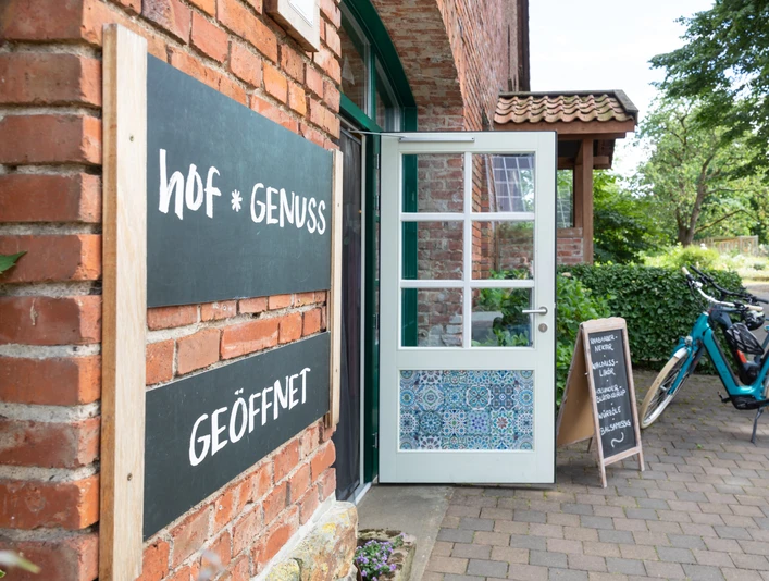 Steinheim-Hagedorn-Hofladen-Teutoburger-Wald-Tourismus-A-Röser-118.jpg Eingang eines Backsteingebäudes mit Schildern "hof * genuss" und "GEÖFFNET", daneben Fahrräder.