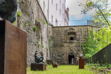 Sculptures on plinths stand in a green courtyard with old stone walls and a building in the background, surrounded by trees.