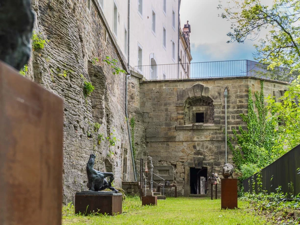 Sculptures on plinths stand in a green courtyard with old stone walls and a building in the background, surrounded by trees.