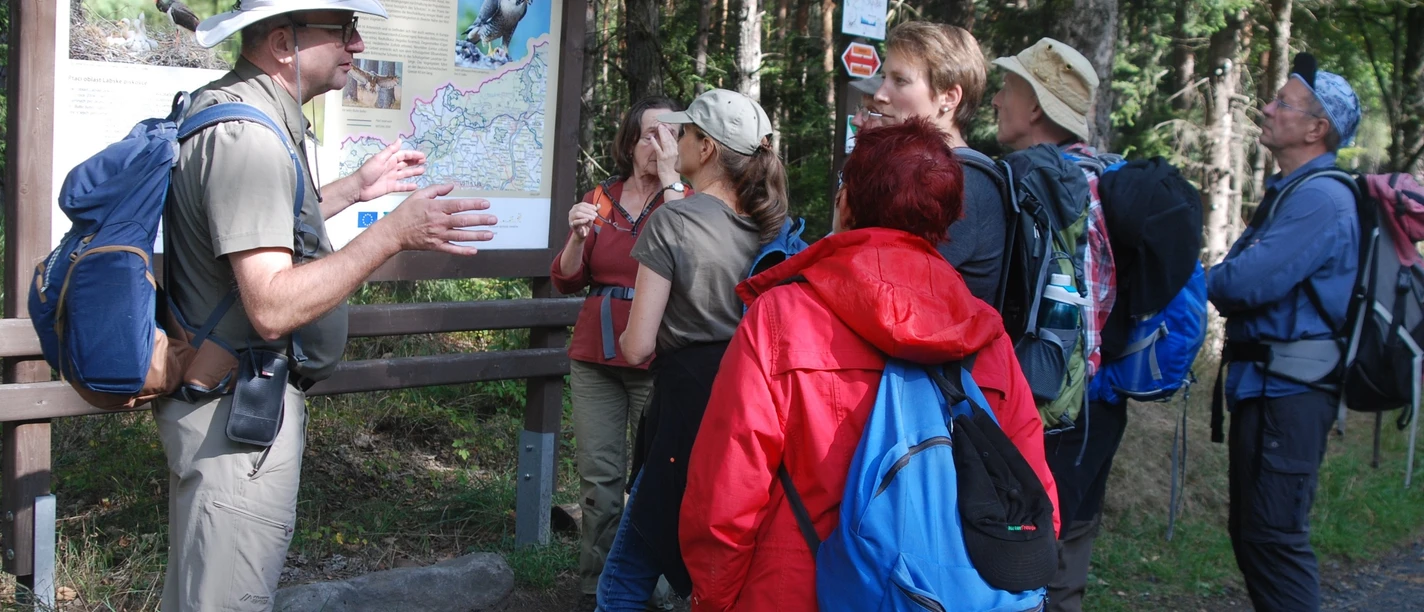 Wandelgids Ralf Schmaedicke Een groep wandelaars staat voor een informatiebord in het bos, een man met een hoed legt iets uit; ze dragen kleurrijke rugzakken.