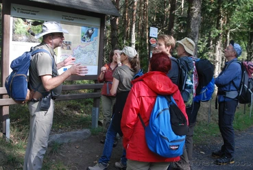 Wanderführer Ralf Schmaedicke Eine Gruppe von Wanderern steht vor einem Infotafel im Wald, ein Mann mit Hut erklärt etwas; sie tragen bunte Rucksäcke.
