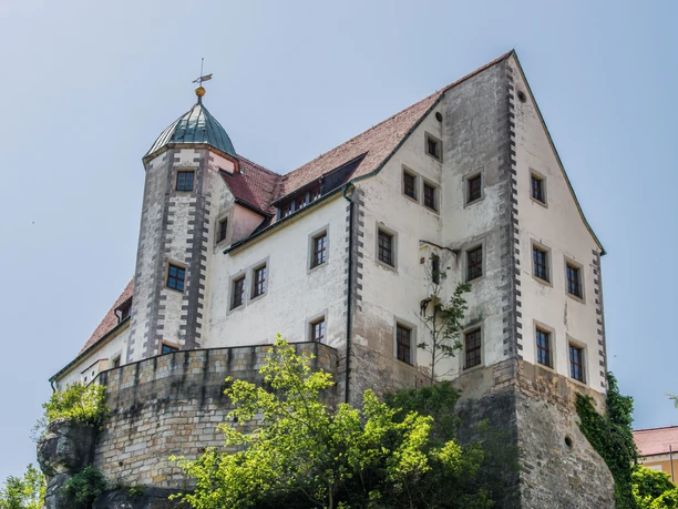 Burg Hohnstein Historisches Schloss mit weißer Fassade und rotem Ziegeldach auf einem Felsen, umgeben von grünen Bäumen, unter klarem, blauem Himmel.