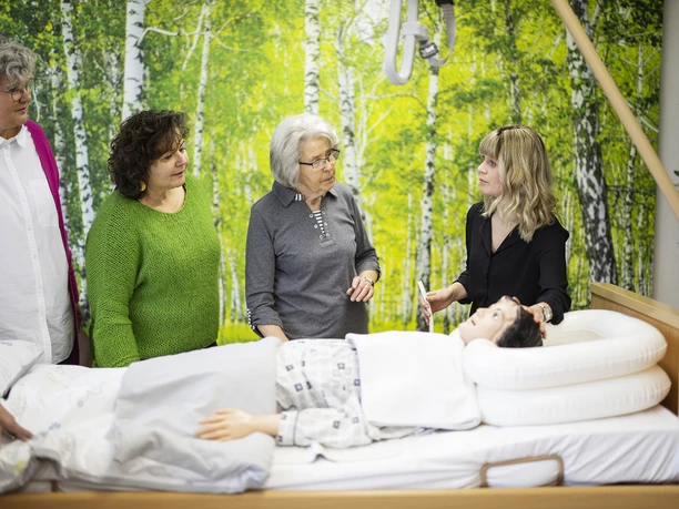 Pflegetraining MZG Bad Lippspringe Frauen diskutieren im Pflegetraining um ein Krankenbett mit Übungspuppe in einer grünen Umgebung.