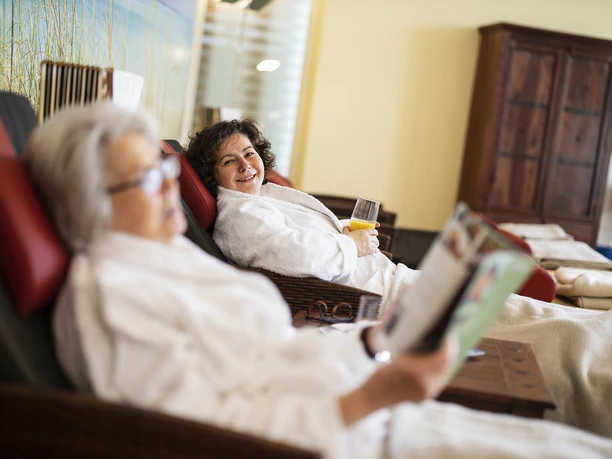 Relaxen MZG Bad Lippspringe Zwei Personen entspannen in weißen Bademänteln auf gepolsterten Liegestühlen, eine hält ein Getränk.