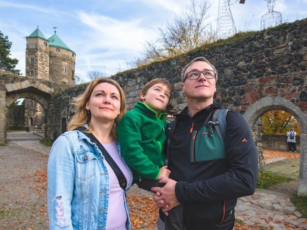Burg Stolpen Eine lächelnde Familie steht vor einer alten Steinmauer der Burg Stolpen mit einem Torbogen, im Hintergrund ein Turm und herbstliche Bäume.