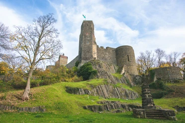 Burg Stolpen Die Burg Stolpen mit hohen, grauen Mauern auf einem grünen Hügel, umgeben von herbstlichen Bäumen unter blauem Himmel.