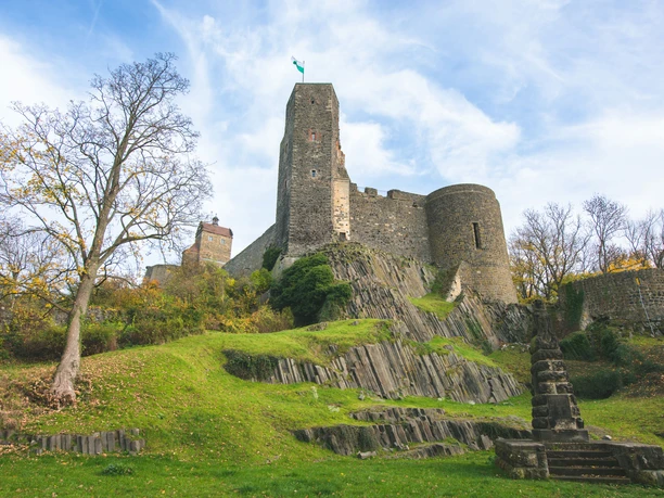 Burg Stolpen Die Burg Stolpen mit hohen, grauen Mauern auf einem grünen Hügel, umgeben von herbstlichen Bäumen unter blauem Himmel.