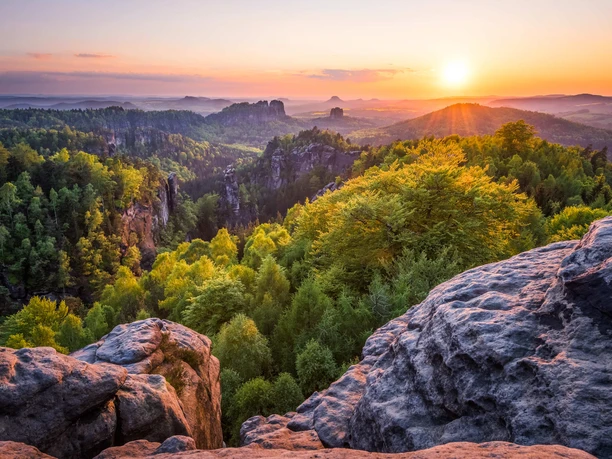 Blick vom Carolafelsen Sonnenuntergang über einer grünen Waldlandschaft mit Felsen im Vordergrund und sanften Hügeln im Hintergrund, warme und friedliche Atmosphäre.