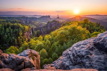 View from Carola Rock Sunset over a green forest landscape with rocks in the foreground and rolling hills in the background, warm and peaceful atmosphere.