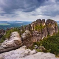 Blick von der Schrammsteinaussicht Felsformationen der Schrammsteine in der Sächsischen Schweiz, umgeben von dichtem Wald unter einem bewölkten Himmel, mit Blick auf eine weite Landschaft.