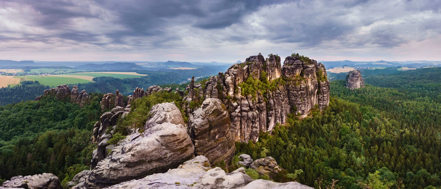 Blick von der Schrammsteinaussicht Felsformationen der Schrammsteine in der Sächsischen Schweiz, umgeben von dichtem Wald unter einem bewölkten Himmel, mit Blick auf eine weite Landschaft.