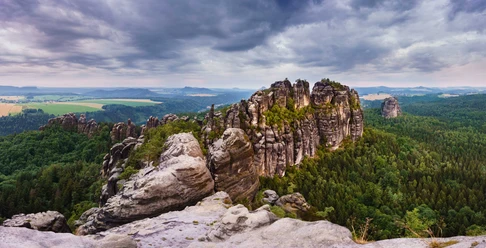 Uitzicht vanaf de Schrammstein uitkijkpost Rotsformaties van de Schrammsteine in Saksisch Zwitserland, omgeven door dichte bossen onder een bewolkte hemel, met uitzicht op een uitgestrekt landschap.