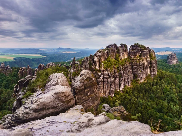 View from the Schrammstein lookout Rock formations of the Schrammsteine in Saxon Switzerland, surrounded by dense forest under a cloudy sky, with a view of a vast landscape.