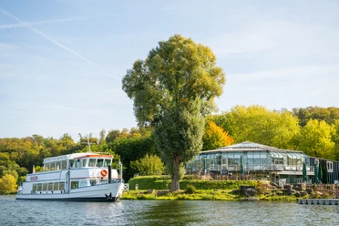 Schieder-Schwalenberg-Schiedersee-Teutoburger-Wald-Tourismus-D-Ketz-074-CC-BY-SA.jpg Weißes Ausflugsschiff auf einem Fluss, umgeben von grünen Bäumen und einem Gewächshaus am Ufer.