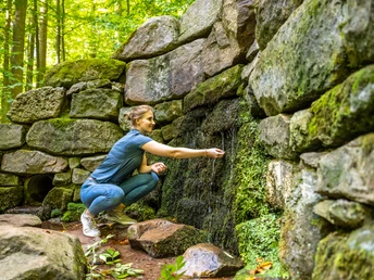 Frau kniet an einer moosbedeckten Steinmauer in einem Wald und berührt sanft einen kleinen Wasserquell.
