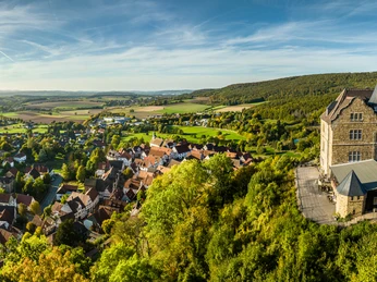 Luftaufnahme eines malerischen Dorfes in hügeliger Landschaft mit historischem Schloss am rechten Bildrand.
