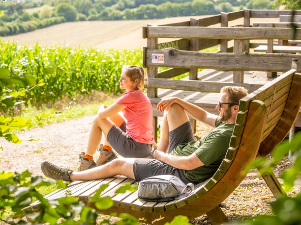 Borgholzhausen-Aussichtsplattform Berghauser Weg-Teutoburger-Wald-Tourismus-D-Ketz-072.jpg Aussichtsplattform mit Holzliegen, zwei entspannten Personen und Blick auf grüne Landschaft im Sommer.
