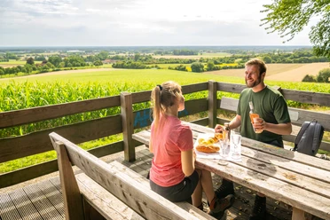 Zwei Personen genießen ein Picknick mit Blick auf weite Felder und Wälder von einer Holzplattform.