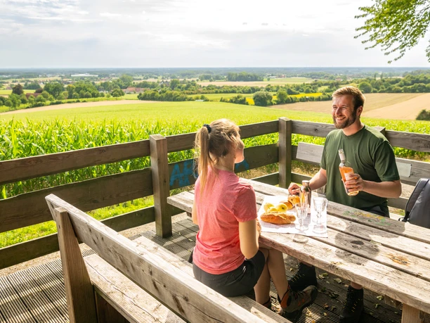 Borgholzhausen-Aussichtsplattform Berghauser Weg-Teutoburger-Wald-Tourismus-D-Ketz-073.jpg Zwei Personen genießen ein Picknick mit Blick auf weite Felder und Wälder von einer Holzplattform.