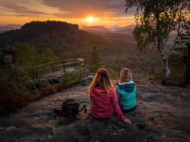 Blick beim Sonnenuntergang vom Papststein Zwei Personen sitzen auf einem Felsen und blicken in einen bewaldeten Sonnenuntergang. Eine trägt eine rote, die andere eine blaue Jacke.