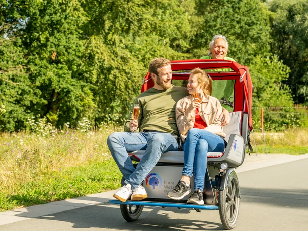 Rheda-Wiedenbrueck-Flora-Westfalica-Park, Rikscha-Fahrt-Teutoburger-Wald-Tourismus-D-Ketz-016.jpg Ein Paar genießt eine gemütliche Rikschafahrt im Park, während der Fahrer lächelnd das Fahrrad steuert.