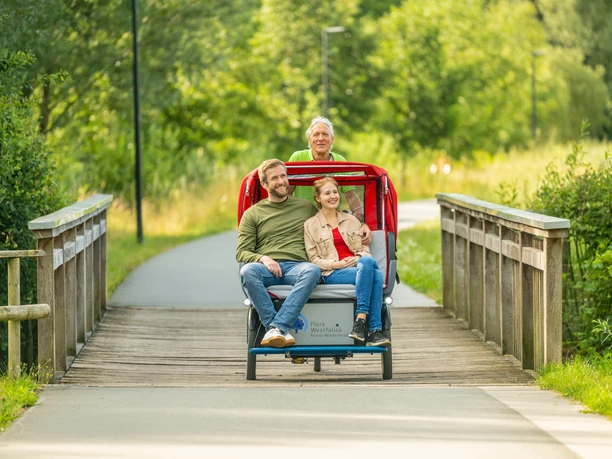 Rheda-Wiedenbrueck-Flora-Westfalica-Park, Rikscha-Fahrt-Teutoburger-Wald-Tourismus-D-Ketz-012.jpg Drei lachende Menschen genießen eine Fahrt mit einer Rikscha auf einem sonnigen Waldweg.