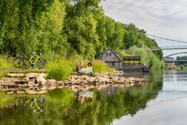 Minden-Schiffmühle-Teutoburger-Wald-Tourismus-Patrick-Gawandtka-021.jpg Personen sitzen entspannt am Flussufer der Weser bei Minden; Fahrräder, eine alte Hütte, und Brücke.
