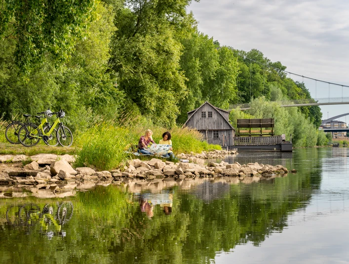 Minden-Schiffmühle-Teutoburger-Wald-Tourismus-Patrick-Gawandtka-021.jpg Personen sitzen entspannt am Flussufer der Weser bei Minden; Fahrräder, eine alte Hütte, und Brücke.