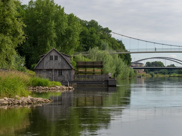 Historische Wassermühle an der Weser bei Minden, umgeben von grünen Bäumen unter grauem Himmel.