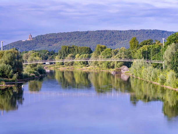 Minden-Schiffmühle-Teutoburger-Wald-Tourismus-Patrick-Gawandtka-006-CC-BY-SA.jpg Ein idyllischer Fluss mit Hängebrücke und bewaldeten Hügeln im Hintergrund, unter blauem Himmel.