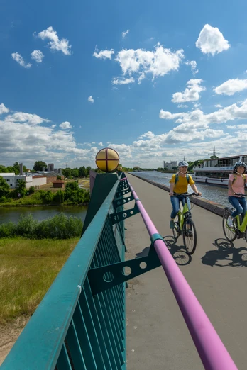 Zwei Radfahrer auf einem breiten Weg entlang eines Flusses, daneben ein vorbeifahrendes Ausflugsschiff.