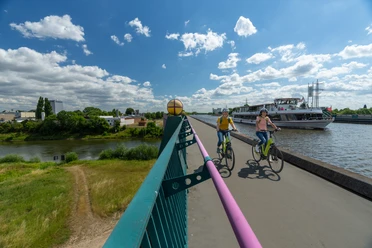 Minden-Wasserstrassenkreuz-Teutoburger-Wald-Tourismus-Patrick-Gawandtka-037.jpg Zwei Radfahrer auf einem breiten Weg entlang eines Flusses, daneben ein vorbeifahrendes Ausflugsschiff.