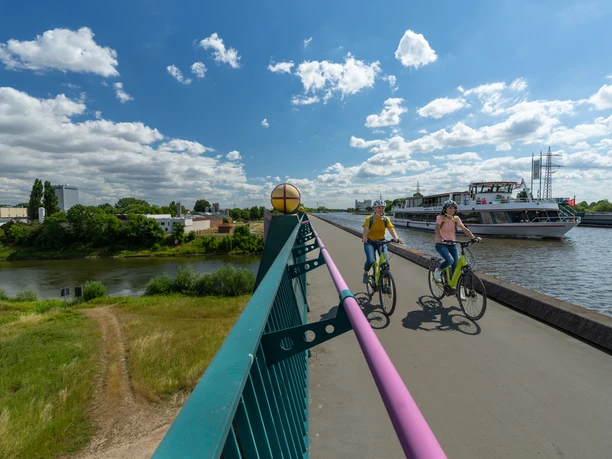 Minden-Wasserstrassenkreuz-Teutoburger-Wald-Tourismus-Patrick-Gawandtka-037.jpg Zwei Radfahrer auf einem breiten Weg entlang eines Flusses, daneben ein vorbeifahrendes Ausflugsschiff.