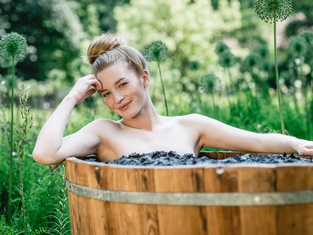 Bad Driburger Moor Junge Frau sitzt in einem Holzfass voller Moor auf einer Wiese, umgeben von grünen Pflanzen.
