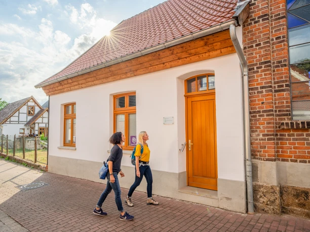 Zwei Frauen gehen auf einem gepflasterten Weg an einem weißen Gebäude mit roten Dachziegeln vorbei.