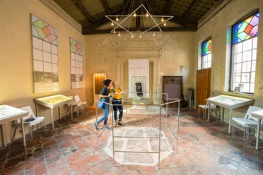 Besucher erkunden eine Ausstellung in einem historischen Raum mit bunten Fenstern und Infotafeln.