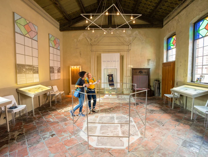 Besucher erkunden eine Ausstellung in einem historischen Raum mit bunten Fenstern und Infotafeln.