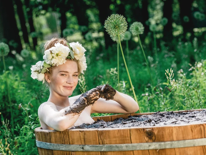 Moorbad Frau mit Blumenkranz im Haar genießt ein Moorbad in hölzerner Wanne, umgeben von Natur.