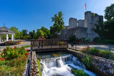Ruine einer Burg mit Flagge im Hintergrund, Brücke über plätschernden Bach, blühende Beete rundum.