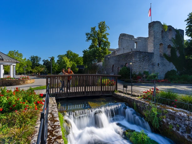 Ruine einer Burg mit Flagge im Hintergrund, Brücke über plätschernden Bach, blühende Beete rundum.